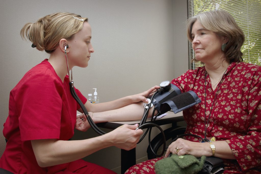 A healthcare professional in red scrubs measuring a female patient's blood pressure, illustrating the ongoing management of pre-existing conditions.