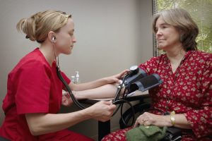A healthcare professional in red scrubs measuring a female patient's blood pressure, illustrating the ongoing management of pre-existing conditions.