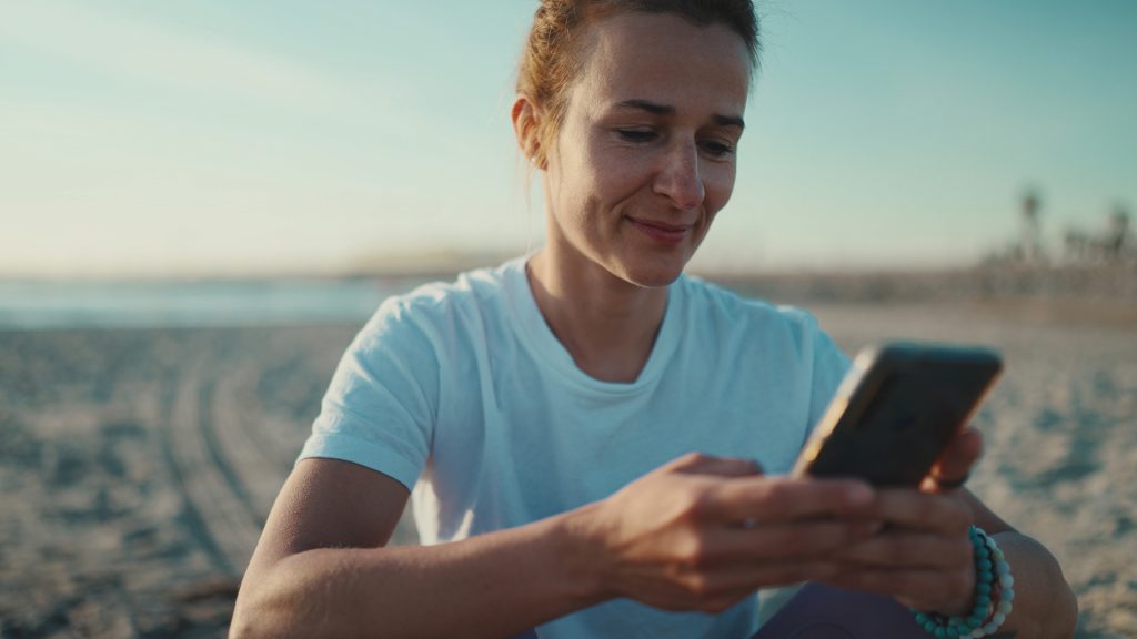 A woman sitting on a beach, looking at her phone while enjoying the view.
