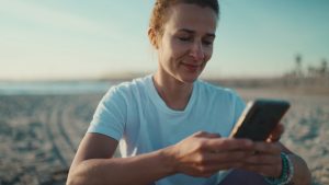 A woman sitting on a beach, looking at her phone while enjoying the view.