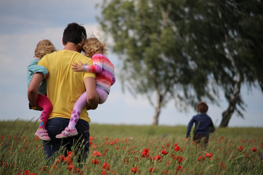 A father carrying two children walks across a wide open field toward a cluster of trees, accompanied by an older child