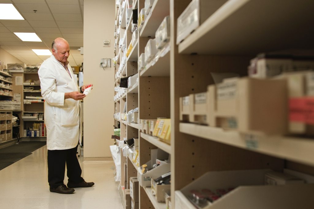 Male pharmacist reading prescription label while surrounded by shelves of medications.